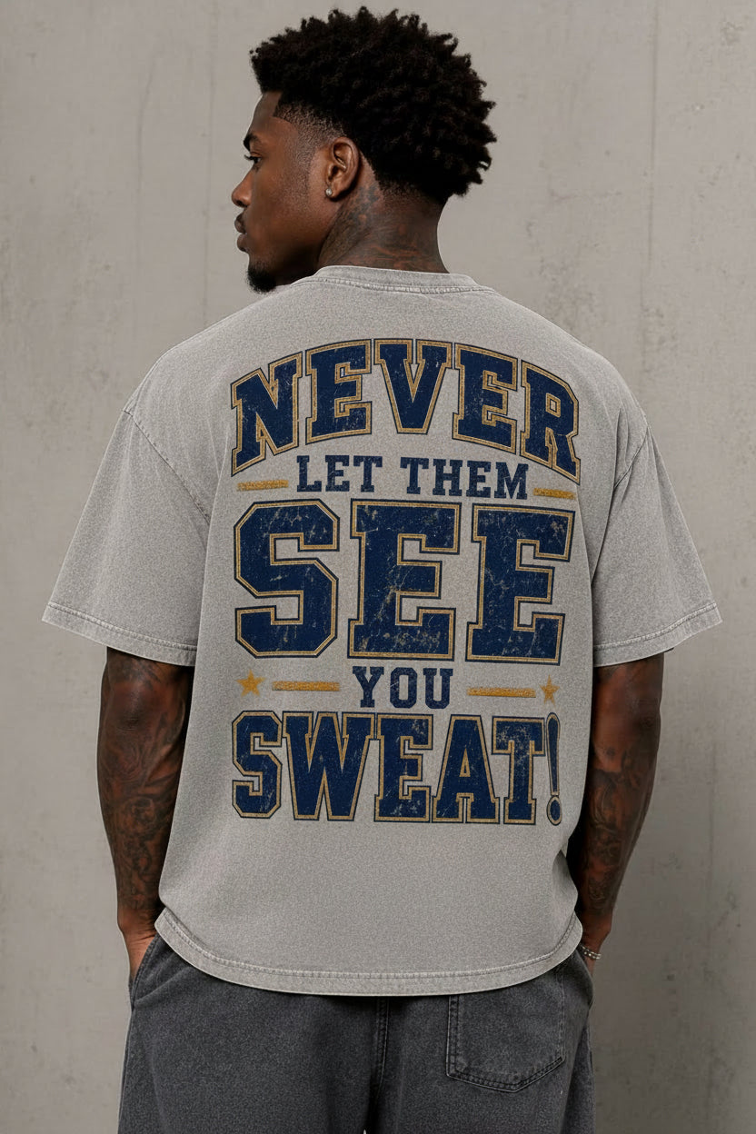 A man viewed from behind wearing a light gray t-shirt with a distressed graphic on the back that reads 'NEVER LET THEM SEE YOU SWEAT!' in navy blue and mustard yellow. He is standing against a textured gray concrete wall.