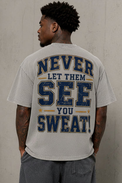 A man viewed from behind wearing a light gray t-shirt with a distressed graphic on the back that reads 'NEVER LET THEM SEE YOU SWEAT!' in navy blue and mustard yellow. He is standing against a textured gray concrete wall.